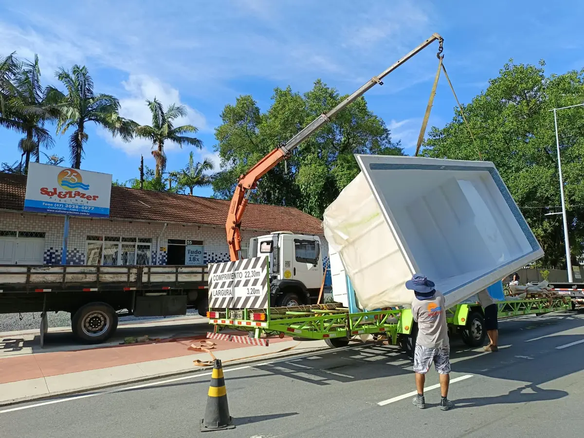 Munck carregando piscina de fibra em carreta na frente de loja de piscinas em Joinville